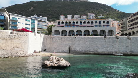 Eine-Wunderschöne-4K-Aufnahme-Einer-Gibraltar-Flagge-Auf-Einem-Felsen-über-Malerischem,-Klarem,-Tropisch-Aussehendem-Wasser-In-Einer-Bucht-In-Den-Gewässern-Gibraltars