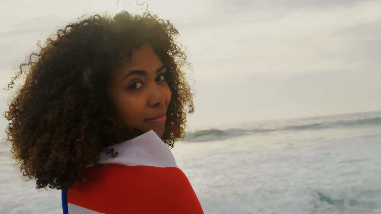 Zijaanzicht van Afro-Amerikaanse vrouw gewikkeld in Amerikaanse vlag staand op het strand 4k