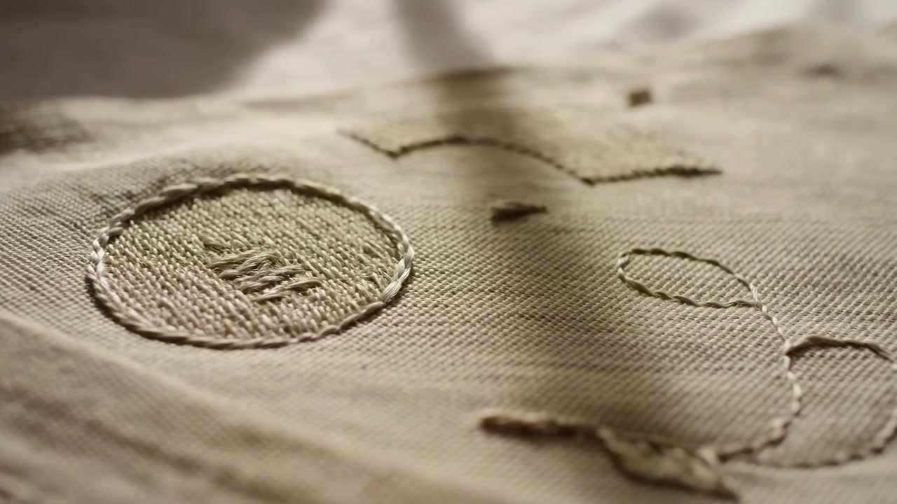 Close view moves gently over neutral woven cloth as stitched circle, scattered thread lines, and loose loop remain in focus, with warm illumination and softly blurred surrounding fabric.