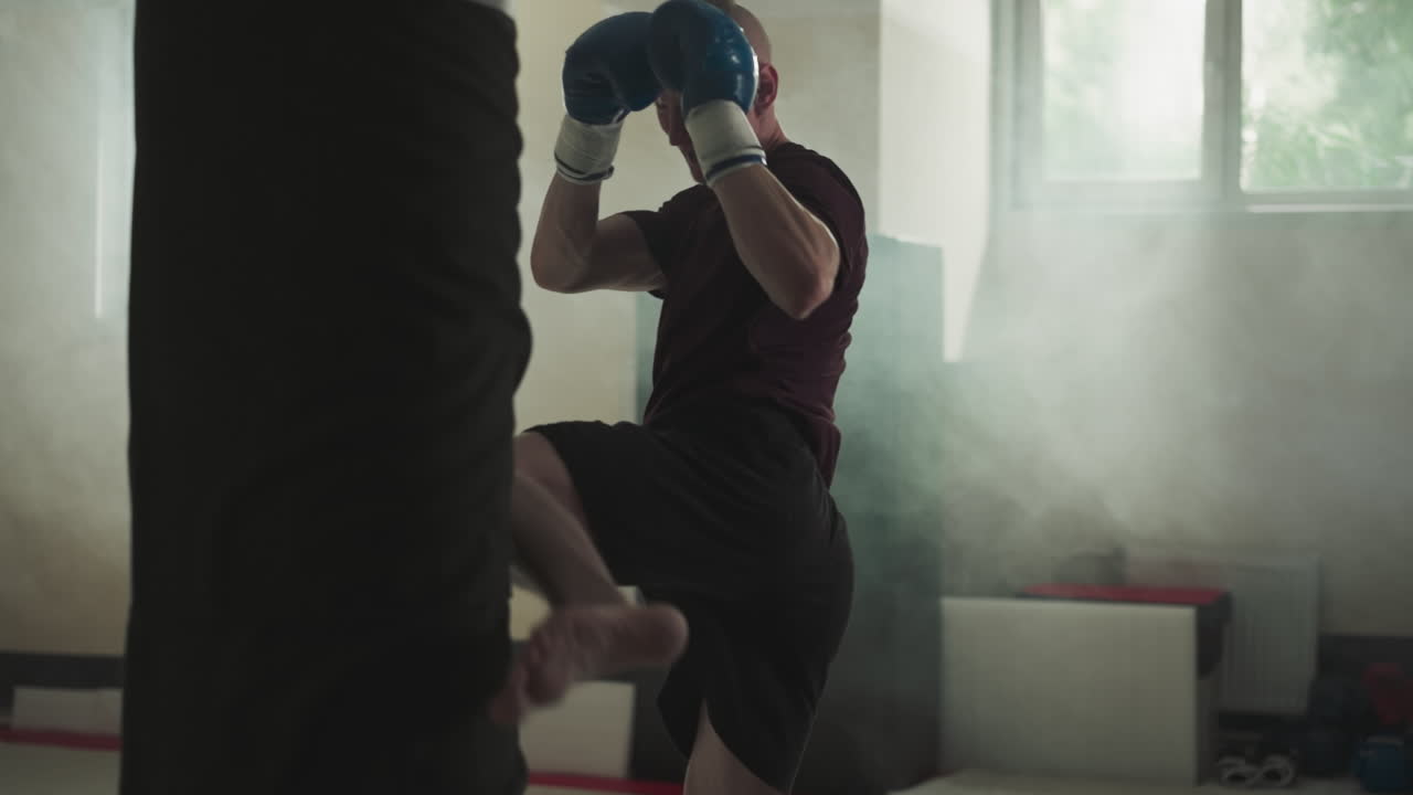 Fighter exercises kicking punching bag in sports center. Concentrated man does roundhouse kick on equipment in foggy gym slow motion. Martial arts