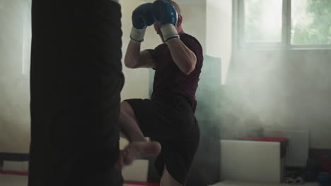 fighter exercises kicking punching bag in sports center. concentrated man does roundhouse kick on equipment in foggy gym slow motion. martial arts