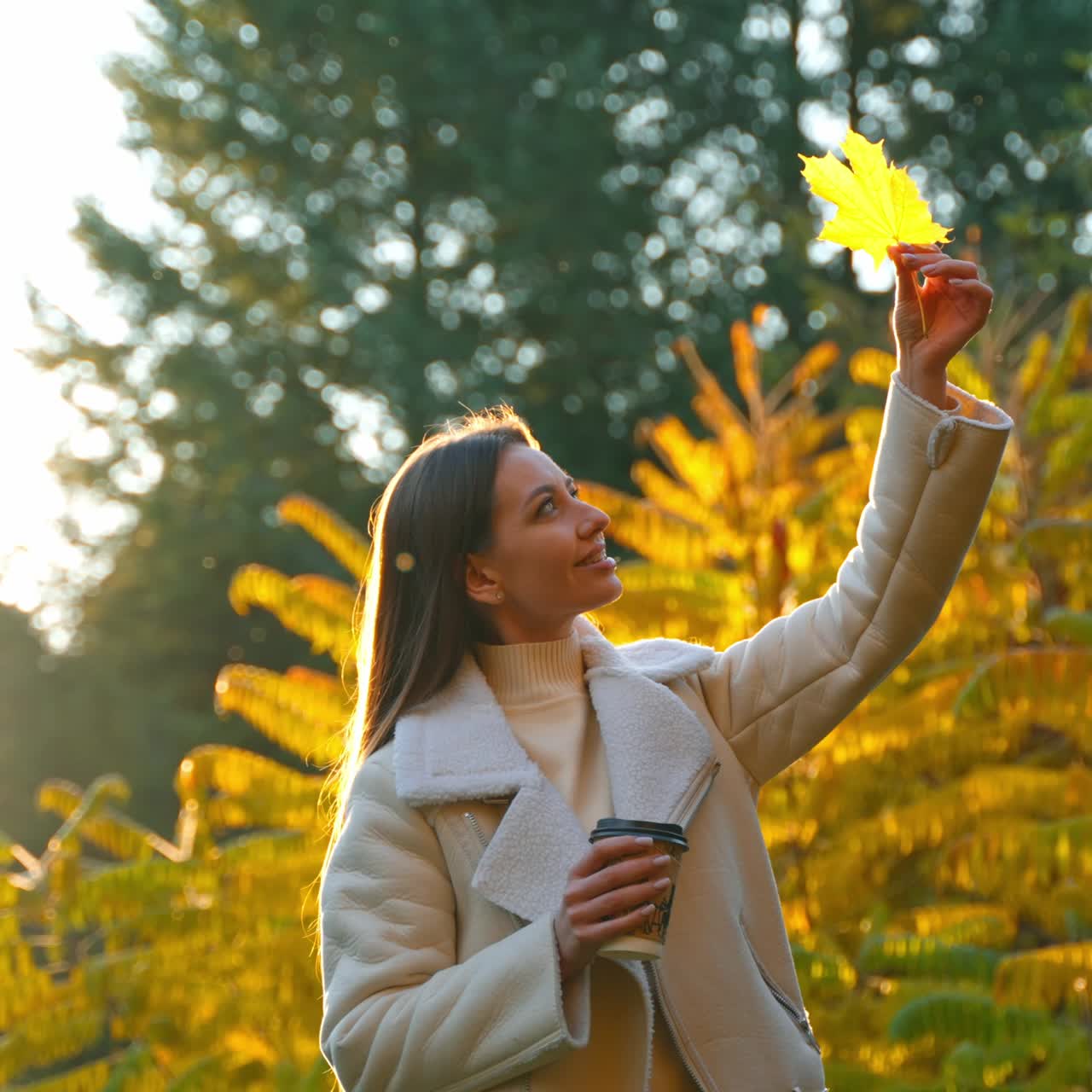 Dark long-haired woman holding yellow maple leaf high above her head. Sunny day in autumn park with a cup of coffee