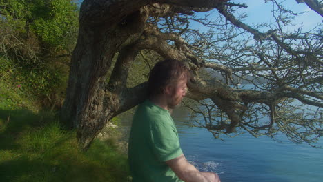 man sitting under shade of lakeside tree on sunny afternoon meditating - close up