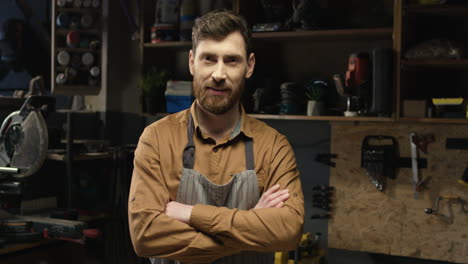 Portrait-shot-of-Caucasian-young-male-carpenter-in-apron-standing-in-workshop-and-smiling-at-camera