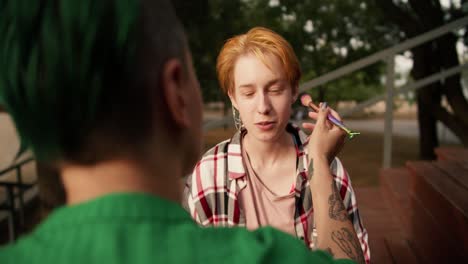 Over-the-shoulder:-a-girl-with-short-green-hair-in-a-green-shirt-doing-makeup-for-her-girlfriend-with-short-orange-hair-in-a-checkered-pink-shirt-on-brown-benches-in-the-park