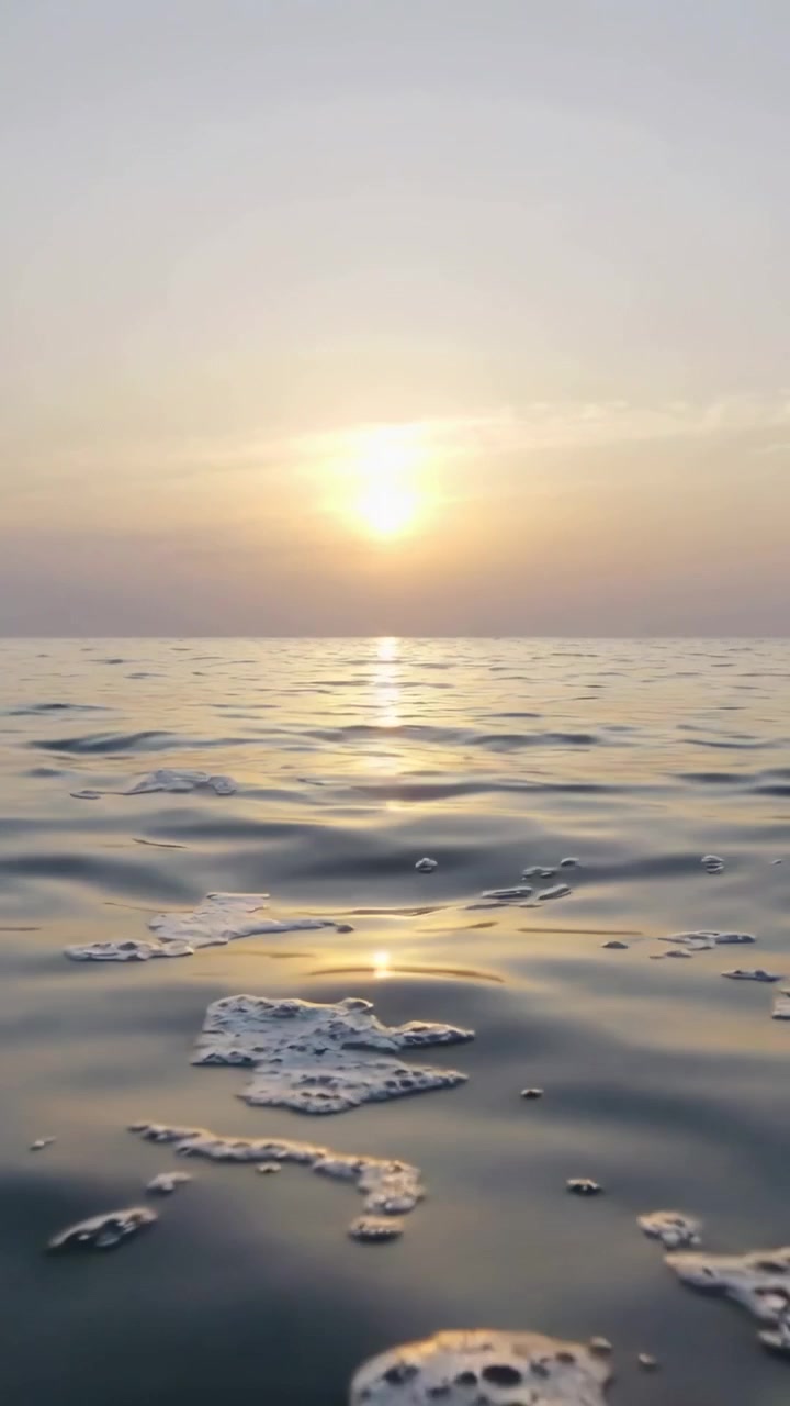 Video verticale: Chiazze di schiuma alla deriva e bagliore del sole vicino all'orizzonte che si muovono sull'acqua, spinte dall'onda lunga.