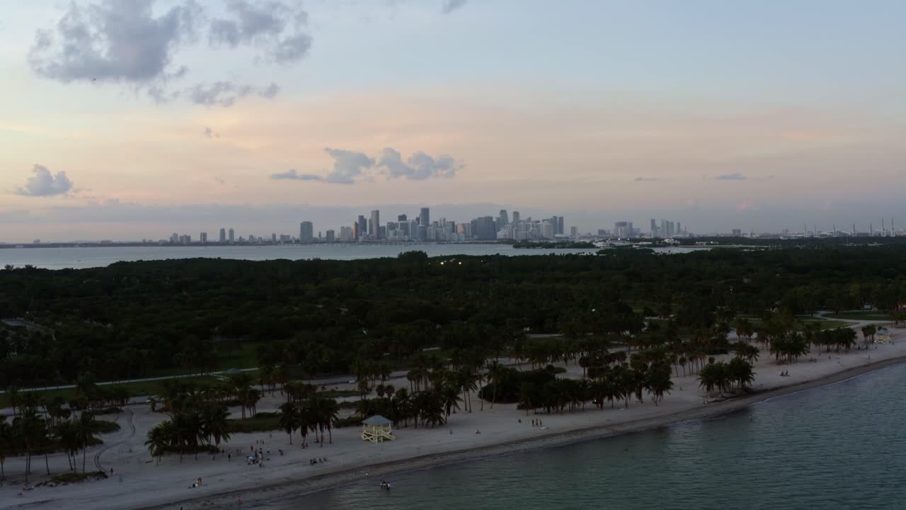 키 비스케인 (key biscayne) 에 있는 크랜던 파크 (crandon park) 에 있는 나무로 둘러싸인 열대 해변의 공중 드론  (aerial drone shot of the tropical beach surrounded by palm trees) 과 플로리다 주 마이애미 (miami) 의 스카이라인 (skyline) 이 먼 곳에서 은 여름 저녁에 보인다.