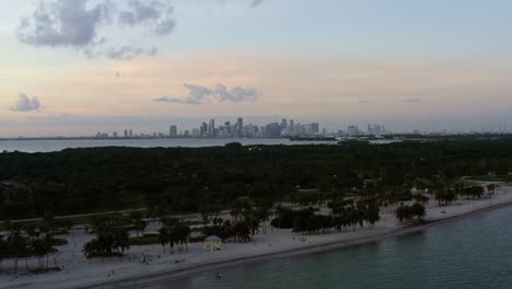 Rising-aerial-drone-shot-of-the-tropical-beach-surrounded-by-palm-trees-on-Crandon-Park-in-Key-Biscayne-with-the-skyline-of-Miami,-Florida-in-the-distance-on-a-sunny-summer-evening