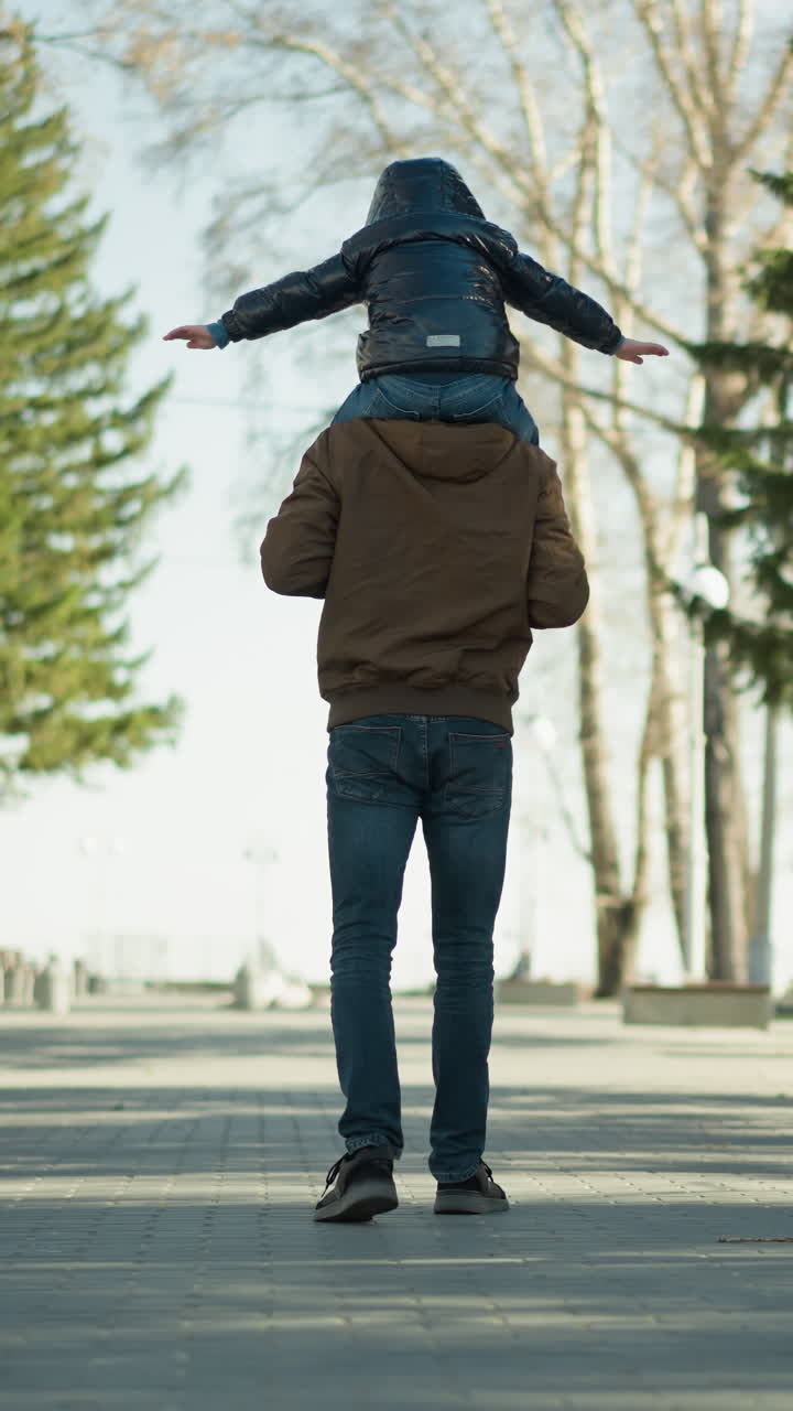 한 아버지가 자신의 아들을 어깨에 고 나무와 전등 기둥으로 둘러싸인 길을 는 동안, 아이는 떨어지는 것을 두려워하는 것처럼 팔을 펴고, 앞으로 나아갑니다.
