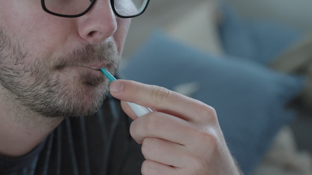 Nahaufnahme von Stock-Videomaterial, das einen Mann zeigt, der seine Körpertemperatur mit einem oralen Thermometer misst, was Fieberkontrolle, Krankheitserkennung, Gesundheitsüberwachung und medizinische Früherkennung zu Hause symbolisiert