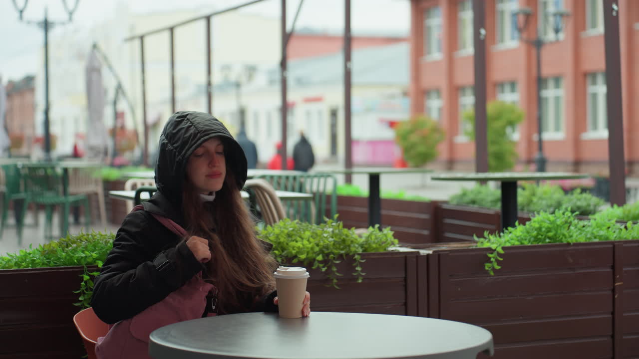 Donna che esce da un bar con una tazza di caffè, cappuccio alzato e borsa rosa a tracolla, che mette un piede sul marciapiede bagnato oltre le vetrine di mattoni, passo deciso e sguardo concentrato, energia da pendolare e tranquilla determinazione