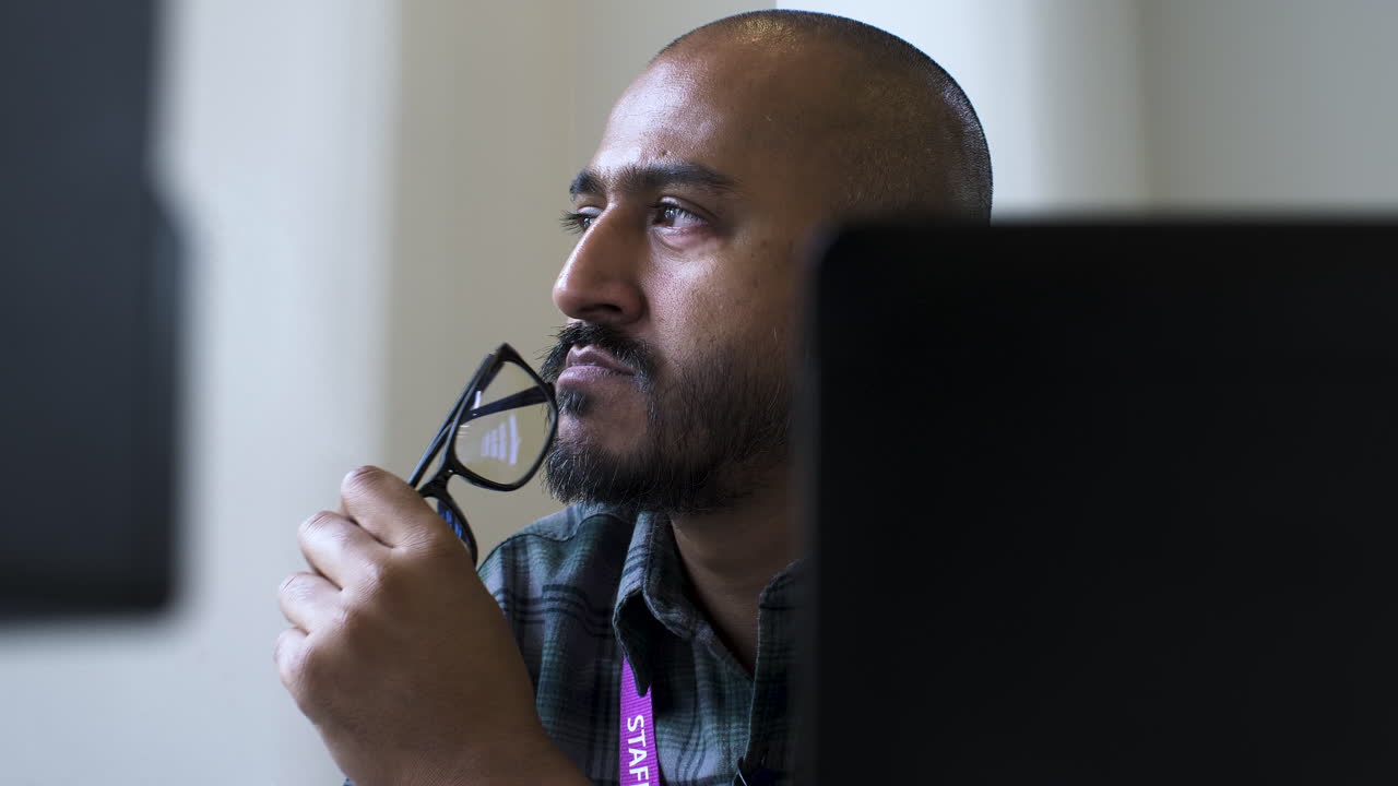 A close up shot from behind an office computer as an Asian man sits gazing into the air in deep thought about an issue while subconsciously tapping his spectacles on his chin