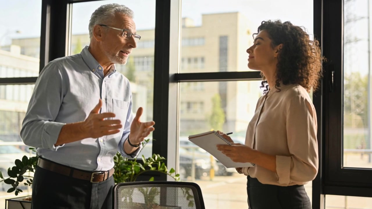 Starting senior manager in shirt clarifying and gesturing while woman holding pad and pen at office