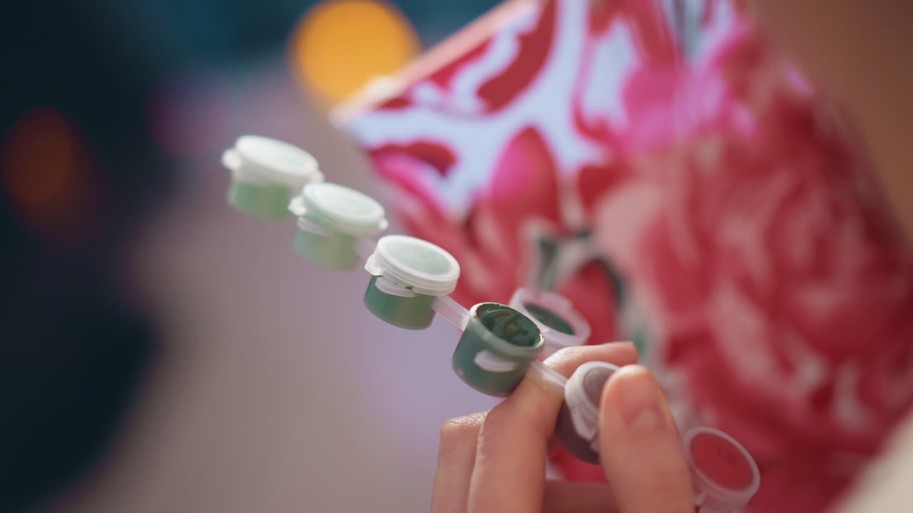 Tight Closeup Of Hands Holding Row Of Small Paint Pots, Caucasian Artist Inspects Lids And Shades Before Applying Color, Neat Palette Arrangement Against Patterned Textile, Studio Selection Moment