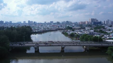 Aerial-fly-in-shot-of-urban-traffic-bridge-and-high-density-waterfront-housing-along-a-canal-in-Ho-Chi-Minh-City,-Vietnam-in-afternoon-light