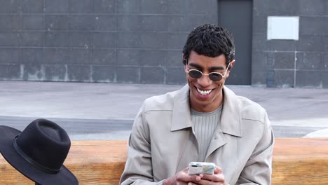 Happy-stylish-ethnic-man-watching-mobile-phone-while-sitting-on-bench