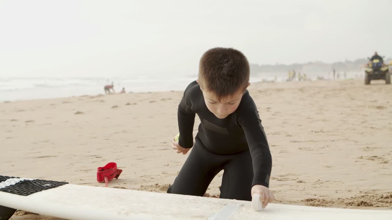 Adorable child waxing surfboard on beach