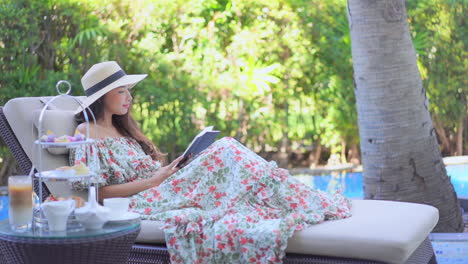 woman in sarafan sundress and hat lying on a deckchair and reading a book outdoors daytime in a tropical resort near the swimming pool