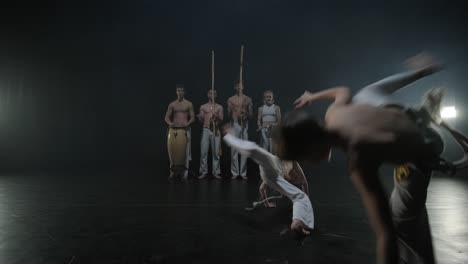 group of a professional dancers are practicing capoeira in darkness against a spotlight on a black background of studio. afro-brazilian martial art that combine elements of dance.