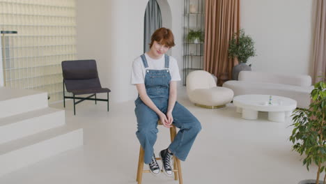 girl in denim jumpsuit sitting on banquette and looking at camera and smiling in the middle of the living room of a modern house