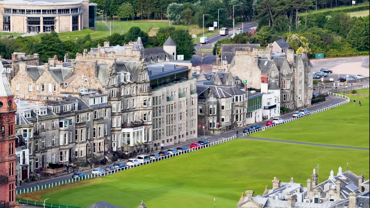 Daytime aerial pan showing historic buildings, green golf course, and parked cars in St Andrews