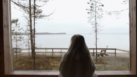 girl is watching the quiet lake landscape outside of the window of her home