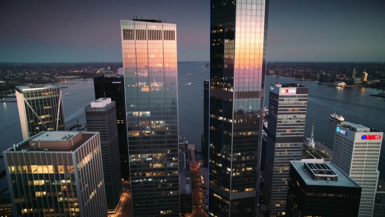 Telecamera aerea che zooma avvicinandosi alla guglia di una torre di vetro al crepuscolo nel porto per catturare il bagliore del tramonto dei traghetti.