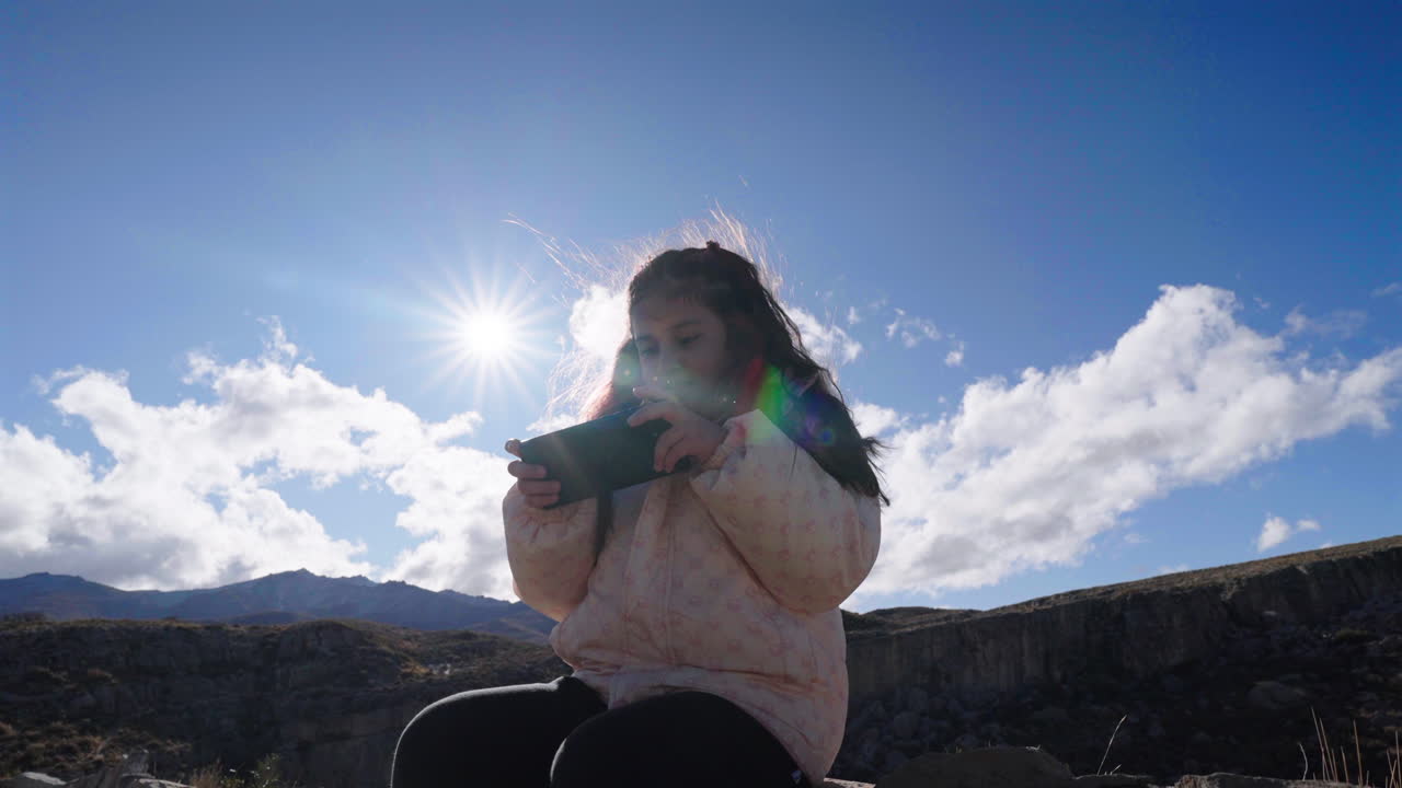Little girl playing mobile video game outdoors under bright sunlight, modern childhood and digital lifestyle