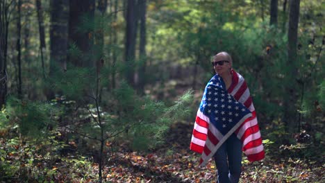 Hübsche-Blonde-Frau,-Die-Durch-Einen-Wald-Geht-Und-Mit-Einer-Amerikanischen-Flagge-Lächelt,-Die-Um-Sie-Gewickelt-Ist