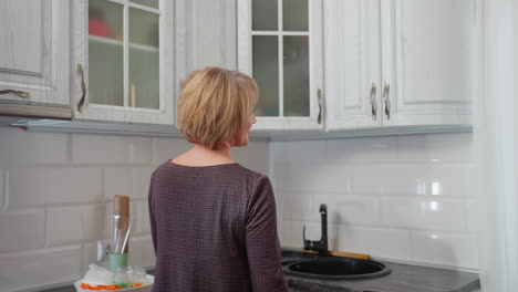 mujer de negocios en túnica entra en su cocina con vista parcial de alguien leyendo un libro, sonríe cálidamente mientras descansa en el armario de la cocina, creando un momento tranquilo y pacífico