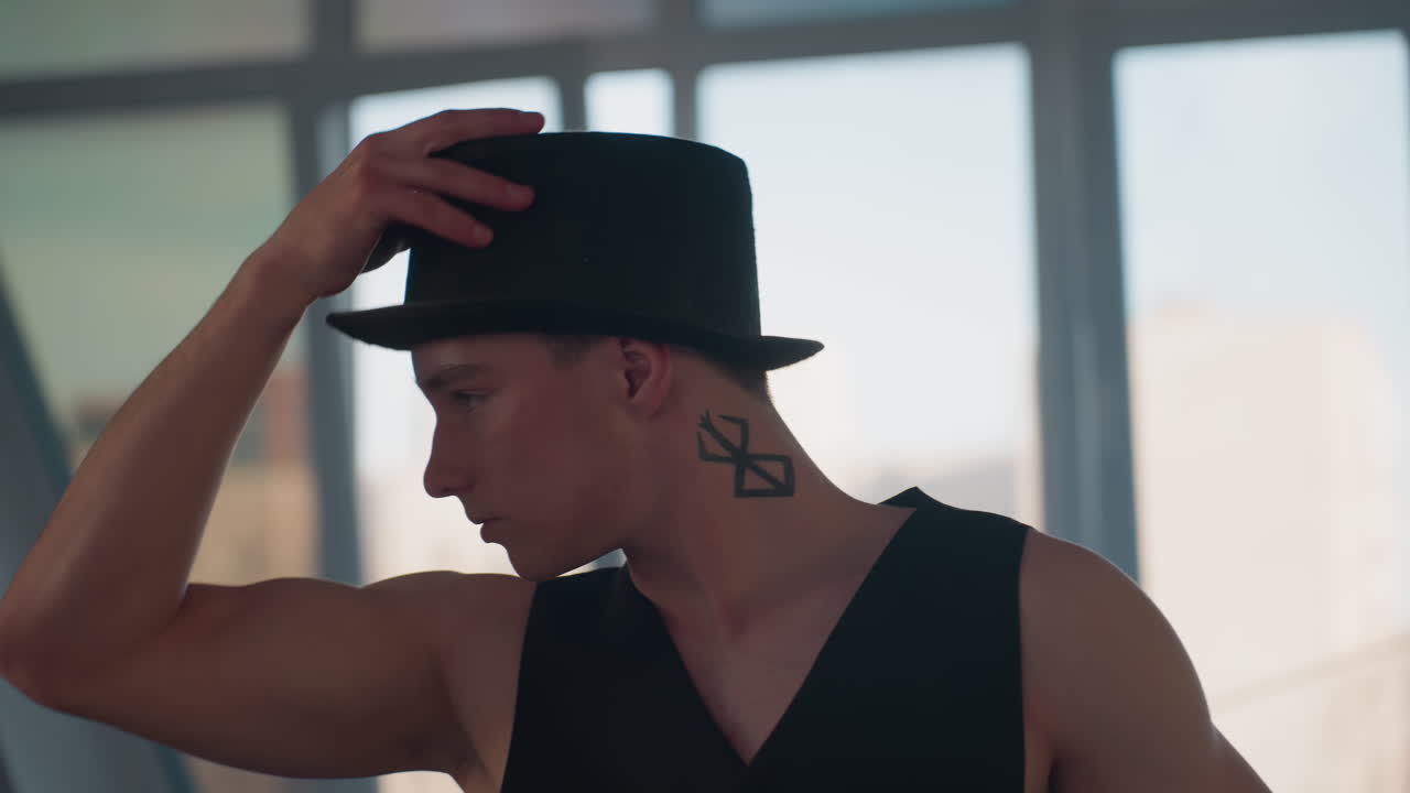 handsome young man dancing in loft studio pauses to remove hat groom hair then replaces hat in stylish vest and pants while smoke and light fill scene mirror reflects movements beside large windows