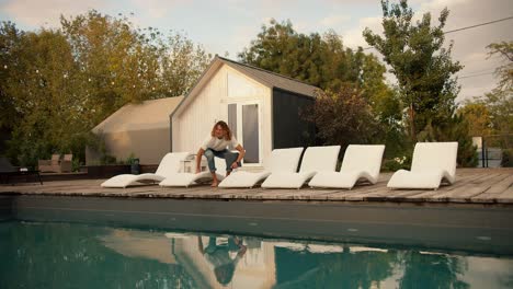 Ein-Typ-Mit-Locken-Und-Brille-Setzt-Sich-Auf-Eine-Weiße-Sonnenliege-Neben-Den-Sonnenliegen-Am-Pool-Und-öffnet-Einen-Laptop.-Ruhepause-Im-Landhaus