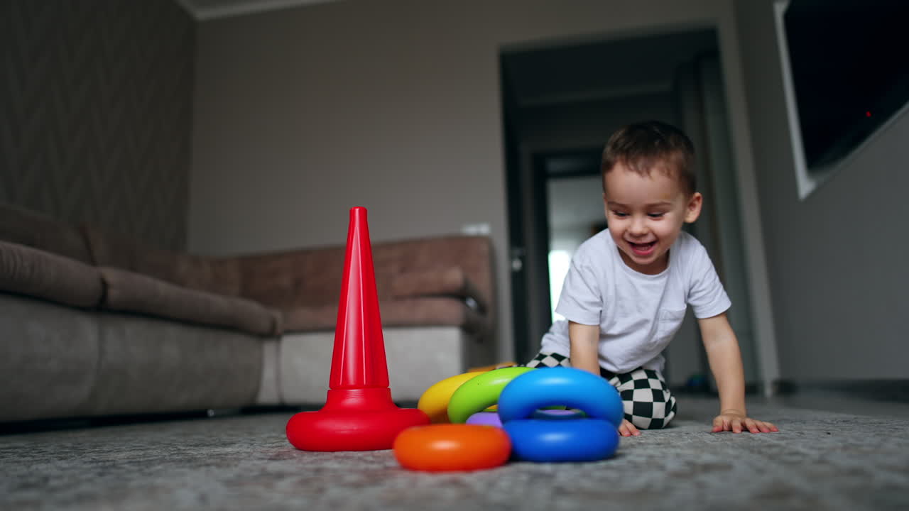 Disassembled toy pyramid lies on the floor. Little baby boy runs up from another room cheerfully and sits down near the toy.
