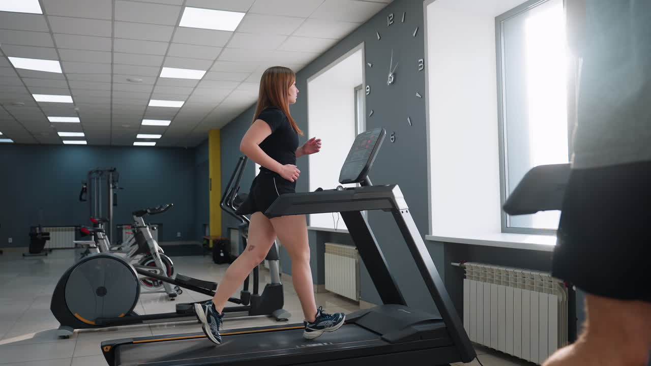 side view of woman running at high speed on treadmill in sunlit modern gym interior with sunlight streaming through windows partial view of another exerciser and cardio equipment on tiled floor