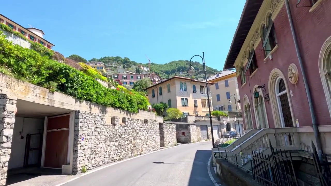 First person perspective driving through narrow winding streets with historic villas and lush green hillsides.