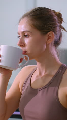 mujer toma un descanso de la sesión de fitness, bebiendo café mientras está sentada en el gimnasio, un gran espejo refleja una postura relajada, equipos de entrenamiento y esteras en el fondo