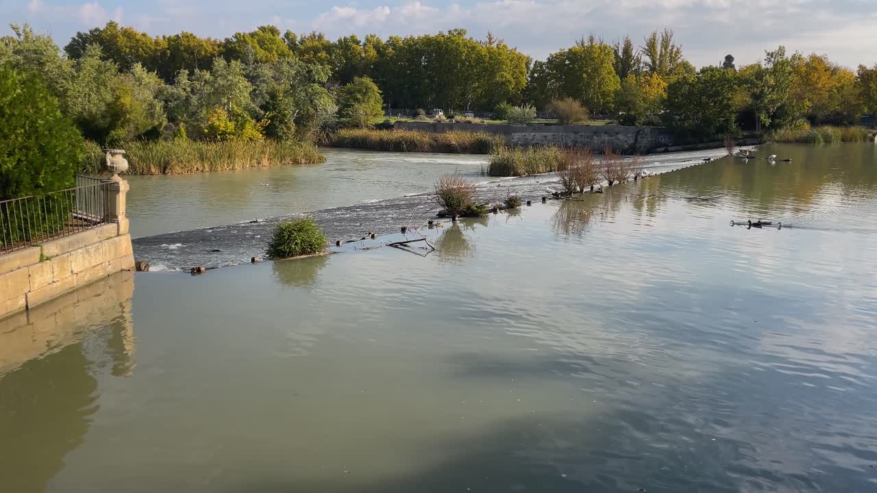 película en cámara lenta de una sección del río tajo a medida que pasa por el palacio real de aranjuez, vemos dos alturas del río con una pendiente donde cae el agua, aparece la balaustrada