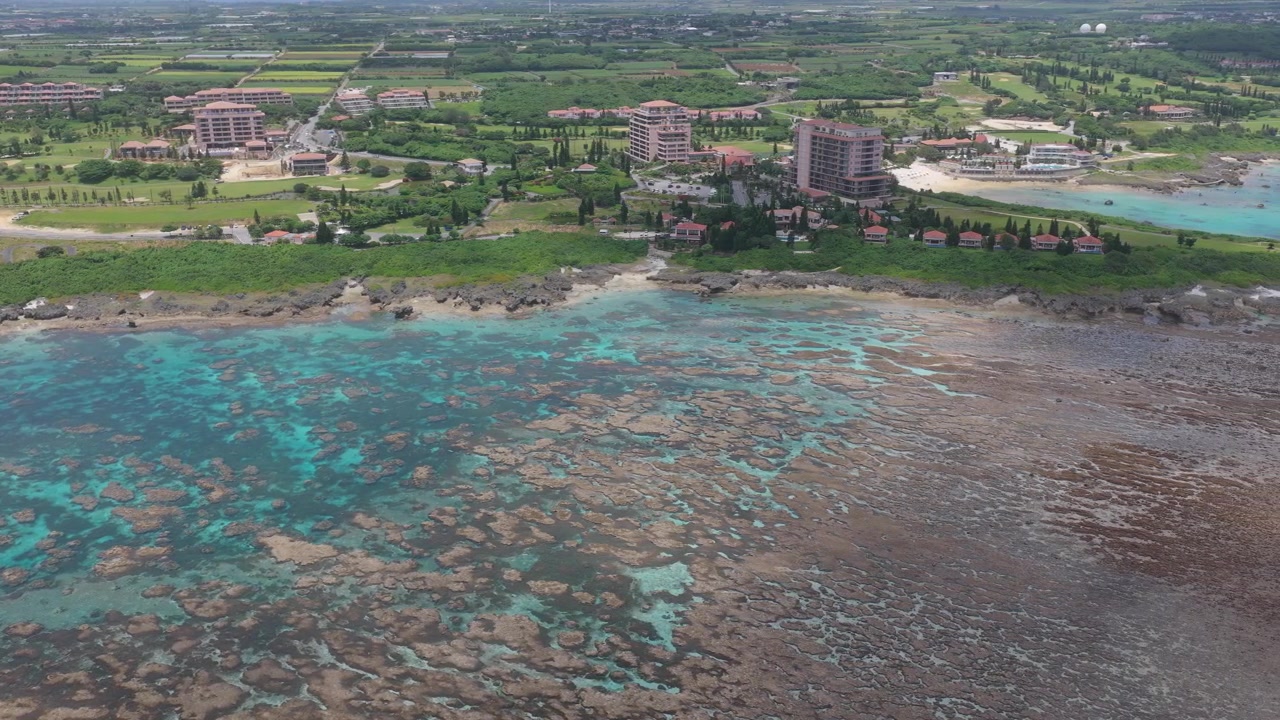 Inquadratura orizzontale con carrellata a destra da un'angolazione obliqua elevata sopra la barriera corallina, acque turchesi e la zona turistica costiera