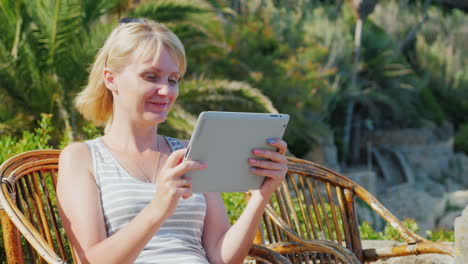 Touristin-Mit-Dem-Tablet-Sagt,-Er-Sitzt-Immer-In-Einem-Korbstuhl-Vor-Dem-Hintergrund-Von-Palmen