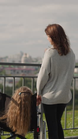 young mother shows city view to daughter with disability standing on viewpoint. little girl sits in wheelchair enjoying spring trip with mom