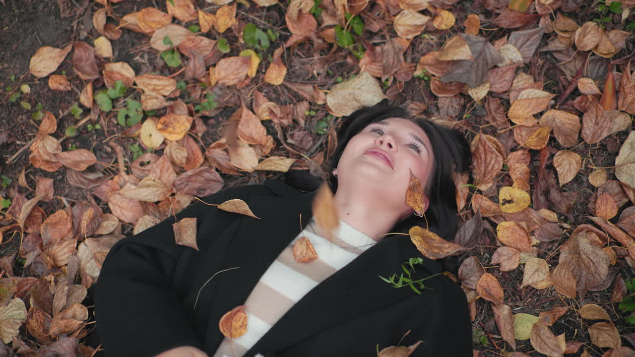 가을 방문객이 숲 바닥에 누워 마른 잎이 얼굴에 떨어지는 동안 미소를 짓고 있어요. 바삭바삭한 갈색 잎과 부드러운 블러 모션으로 둘러싸여 평화로운 야외 분위기, 포근한 검은색 코트와 줄무늬 스웨터가 돋보여요.