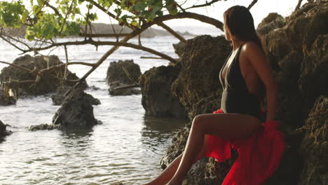 Modelo-De-Chica-Latina-Con-Bronceado-Dorado-Sentado-En-Las-Rocas-De-La-Playa-Tropical-Al-Amanecer-Disfruta-De-La-Vista,-Espacio-De-Copia