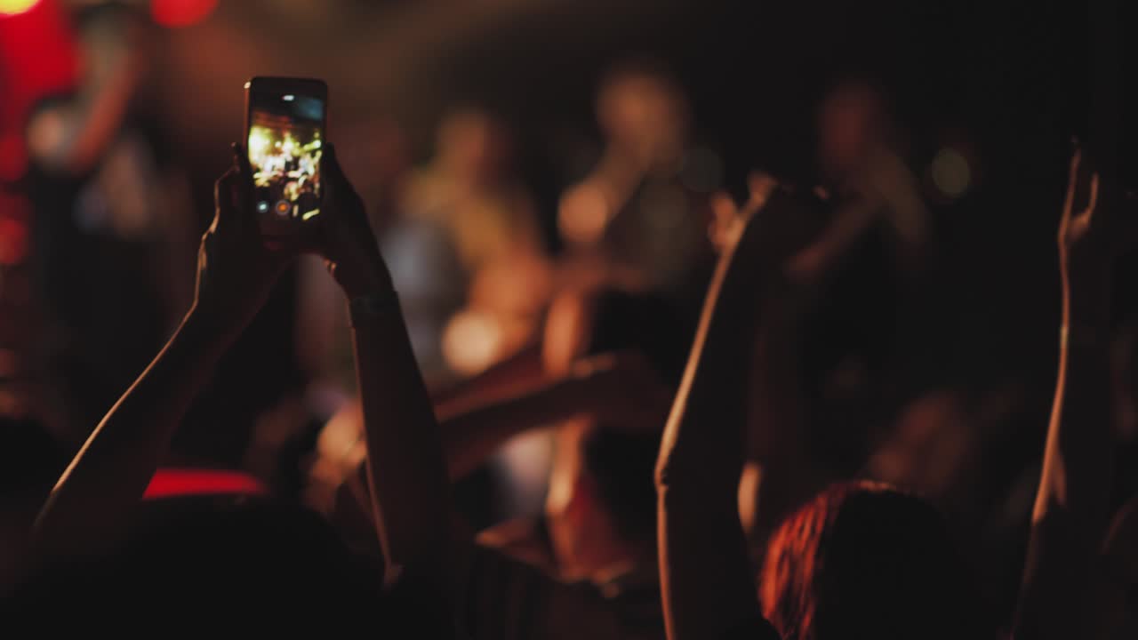 Hands in the crowd of unrecognizable people holding smart phone and shooting musical performance. Bright illumination on the stage