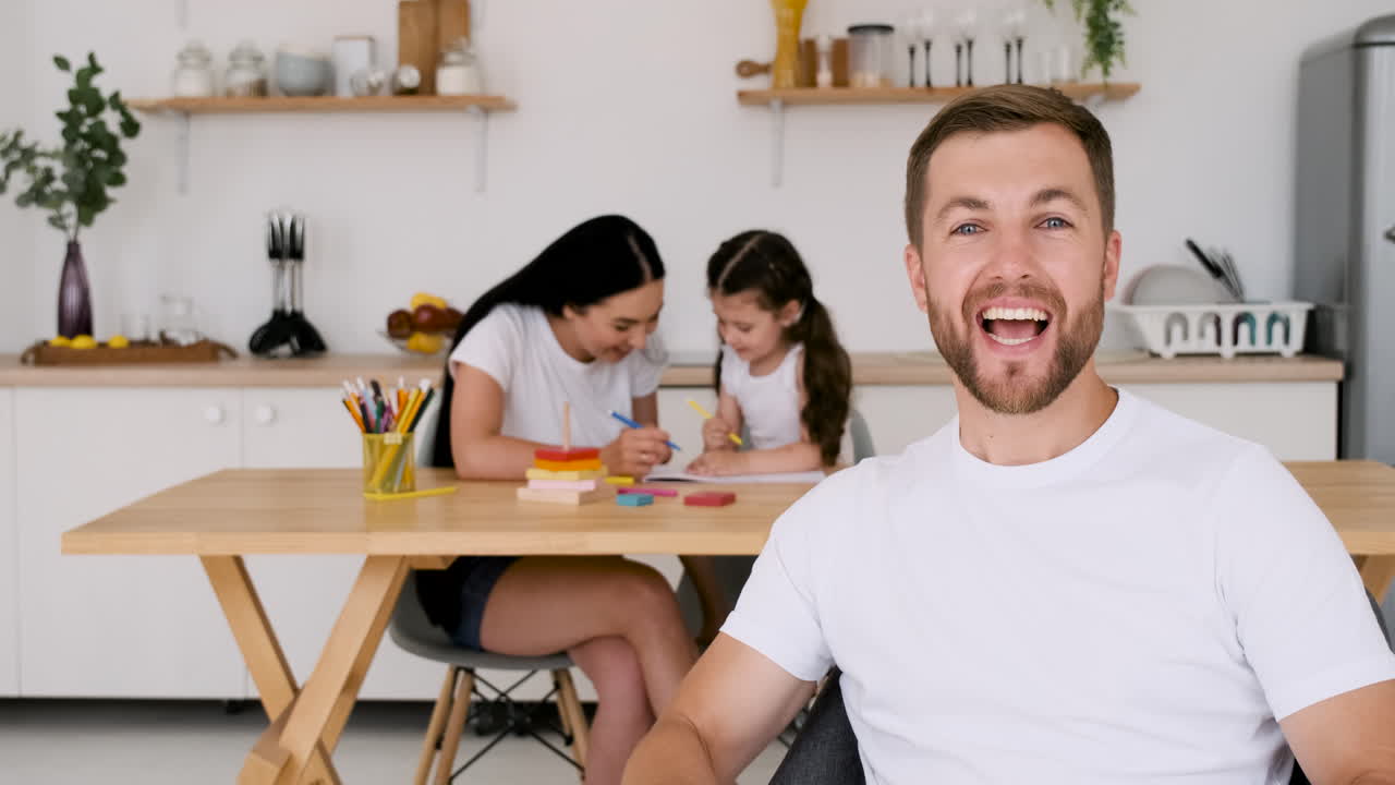 Happy Handsome Man Looking At Camera During A Video Call At Home While On Background His Wife And Little Daughter Sitting At Table And Drawing Together