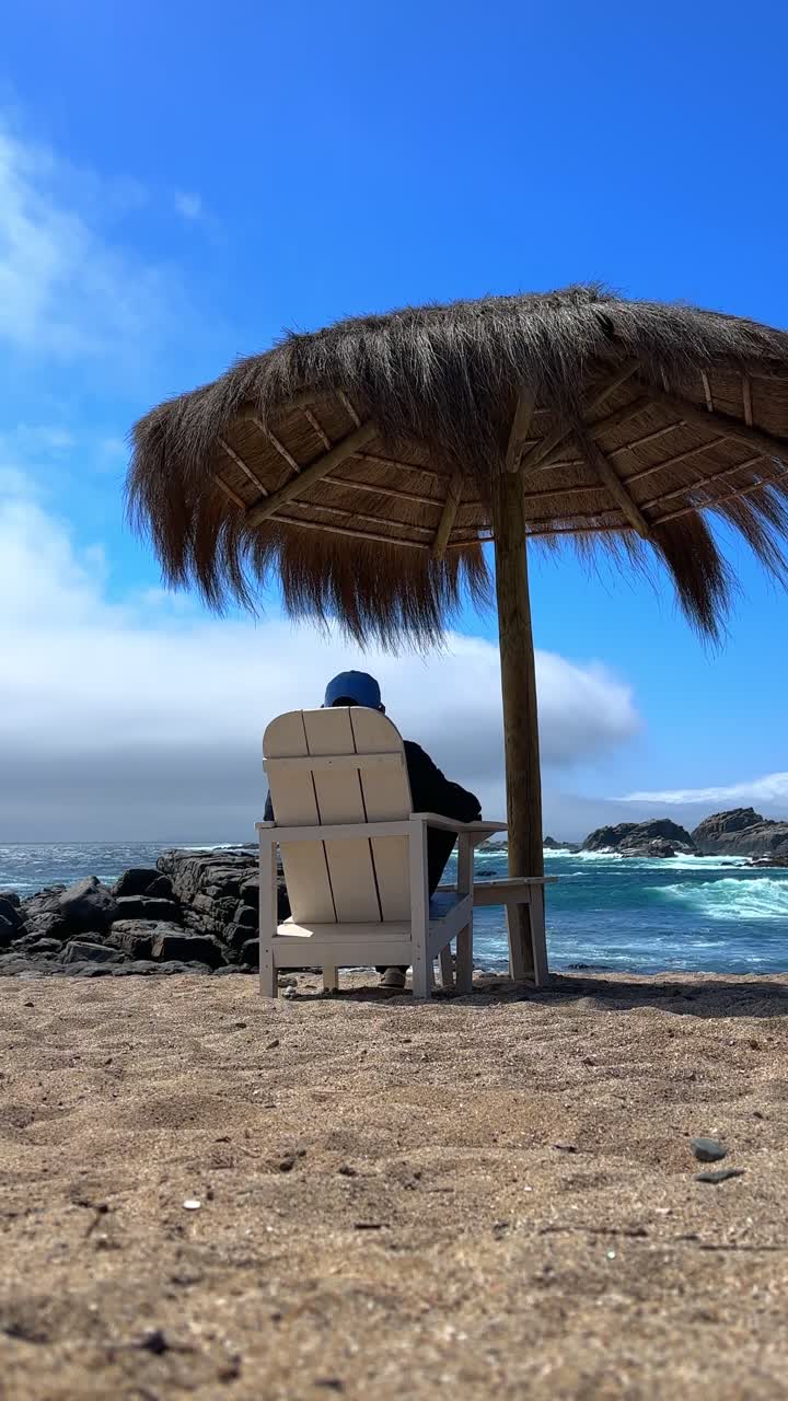 Una ripresa verticale che inquadra un giovane uomo con un berretto, seduto su una sedia di legno sotto un ombrellone fatto di materiali naturali sulla costa della spiaggia di Punta Pite, Cile.