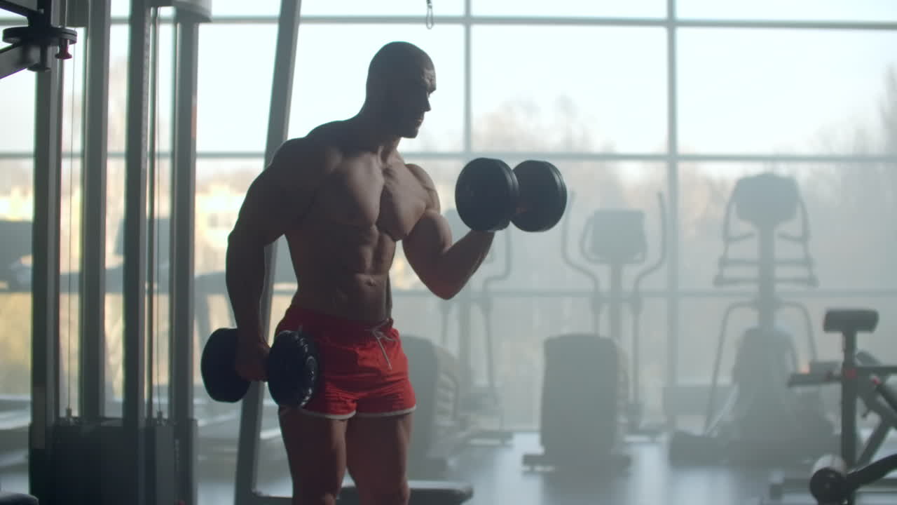 A strong handsome man lifts dumbbells holding them in his hands performing biceps exercises standing at a large window in the GYM modern. Stylish fitness room for training.