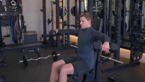 athletic blond man trains his shoulders, doing dumbbell raises to the sides or shoulder fly exercise seated on bench in gym in slow motion