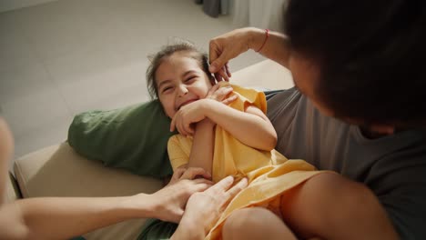 Close-up-shot-of-a-happy-little-girl-in-a-yellow-dress-laughing-and-rejoicing-while-being-tickled-by-her-parents-sitting-on-a-light-brown-in-a-modern-apartment