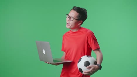 hombre viendo un juego de fútbol en una computadora portátil sosteniendo una pelota de fútbol