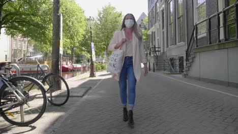 adult blonde lady with face mask walks on brick pavement in amsterdam, netherlands during covid19 outbreak